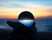 A hand holds a glass sphere which shows the seascape upside down as the sun dips behind the ocean