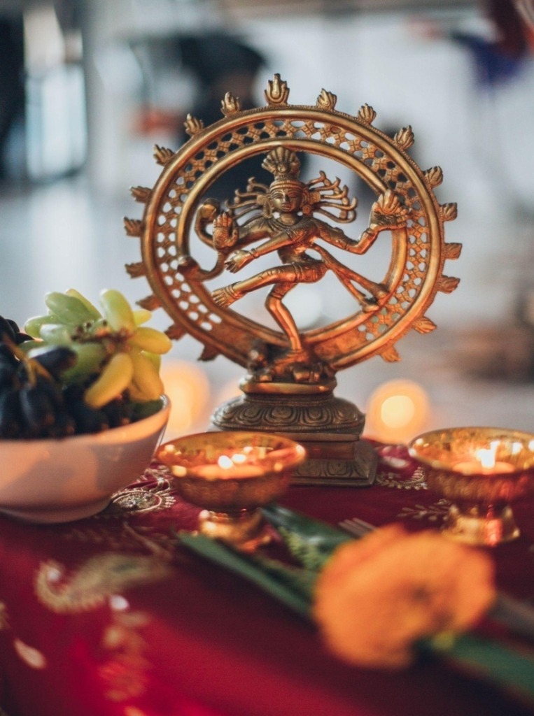 A small idol of Shiva Nataraja surrounded by yellow lit tealights with a bowl of fruit on the left.